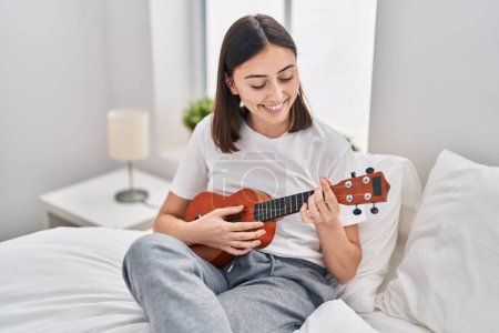 Téléchargez les photos : Young hispanic woman playing ukelele sitting on bed at bedroom - en image libre de droit
