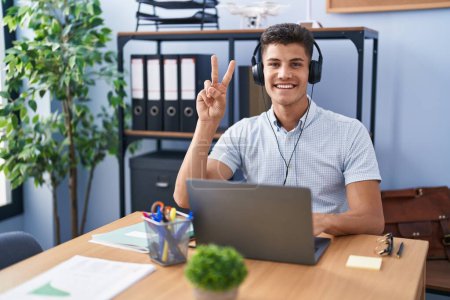 Foto de Joven hombre hispano trabajando en la oficina con auriculares mostrando y señalando con los dedos número dos mientras sonríe confiado y feliz. - Imagen libre de derechos
