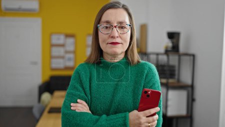 Foto de Mujer hispana madura con trabajador de negocios de cabello gris con cara seria sosteniendo teléfono inteligente en la oficina - Imagen libre de derechos