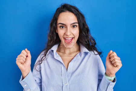 Photo for Young brunette woman standing over blue background celebrating surprised and amazed for success with arms raised and open eyes. winner concept. - Royalty Free Image