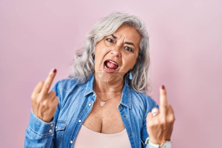Foto de Middle age woman with grey hair standing over pink background showing middle finger doing fuck you bad expression, provocation and rude attitude. screaming excited - Imagen libre de derechos