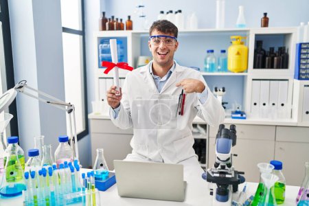 Téléchargez les photos : Jeune homme hispanique travaillant au laboratoire scientifique titulaire d'un diplôme pointant du doigt un sourire heureux et fier - en image libre de droit