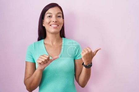 Foto de Mujer hispana joven de pie sobre fondo rosa apuntando hacia atrás con la mano y los pulgares hacia arriba, sonriendo confiada - Imagen libre de derechos