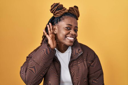 Photo for African woman with braided hair standing over yellow background smiling with hand over ear listening an hearing to rumor or gossip. deafness concept. - Royalty Free Image