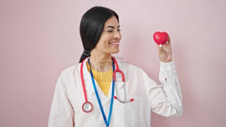 Photo for Young beautiful hispanic woman doctor smiling confident holding heart over isolated pink background - Royalty Free Image