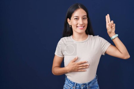 Téléchargez les photos : Jeune femme hispanique debout sur fond bleu souriant jurer avec la main sur la poitrine et les doigts vers le haut, faire un serment de fidélité promesse - en image libre de droit