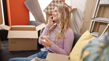 Téléchargez les photos : Jeune femme blonde boire un verre de vin parler sur smartphone à la nouvelle maison - en image libre de droit