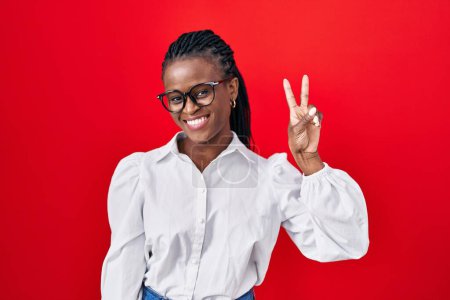 Téléchargez les photos : Femme africaine avec des tresses debout sur fond rouge souriant en regardant la caméra montrant des doigts faisant signe de victoire. numéro deux. - en image libre de droit