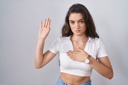Téléchargez les photos : Jeune adolescente debout sur fond blanc jurant avec la main sur la poitrine et la paume ouverte, faisant un serment de fidélité - en image libre de droit