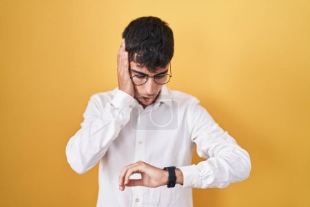 Téléchargez les photos : Jeune homme hispanique debout sur fond jaune regardant le temps de la montre inquiet, peur de se faire tard - en image libre de droit