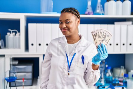 Photo for African american woman with braids working at scientist laboratory holding money looking positive and happy standing and smiling with a confident smile showing teeth - Royalty Free Image