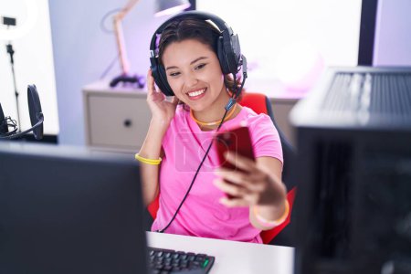 Foto de Mujer hispana joven streamer sonriendo confiado hacer selfie por teléfono inteligente en la sala de juegos - Imagen libre de derechos