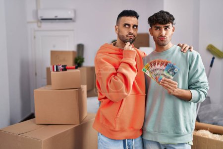 Photo for Young hispanic gay couple moving to a new home holding banknotes serious face thinking about question with hand on chin, thoughtful about confusing idea - Royalty Free Image