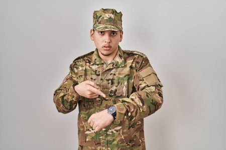 Téléchargez les photos : Jeune homme arabe portant un uniforme de camouflage de l'armée à la hâte pointant pour regarder le temps, l'impatience, bouleversé et en colère pour retard de délai - en image libre de droit