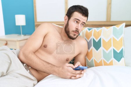 Téléchargez les photos : Jeune homme hispanique utilisant smartphone couché sur le lit dans la chambre - en image libre de droit