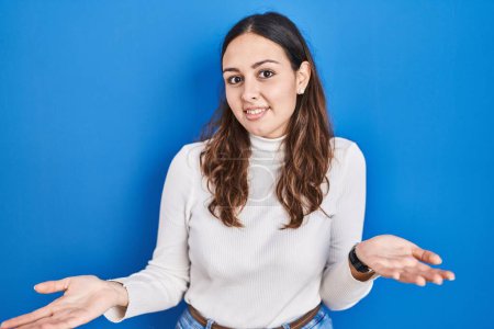 Photo for Young hispanic woman standing over blue background clueless and confused with open arms, no idea concept. - Royalty Free Image