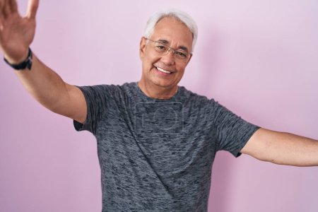 Photo for Middle age man with grey hair standing over pink background looking at the camera smiling with open arms for hug. cheerful expression embracing happiness. - Royalty Free Image