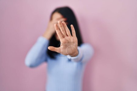 Foto de Mujer hispana de pie sobre fondo rosa cubriendo los ojos con las manos y haciendo un gesto de stop con expresión triste y de miedo. concepto avergonzado y negativo. - Imagen libre de derechos