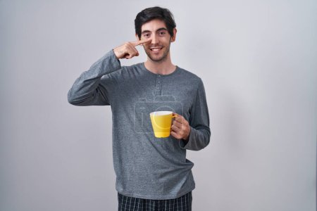 Photo for Young hispanic man wearing pajama drinking a cup of coffee pointing with hand finger to face and nose, smiling cheerful. beauty concept - Royalty Free Image