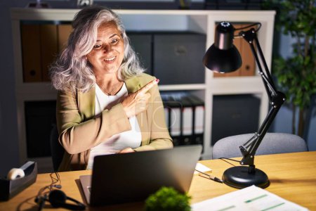 Photo for Middle age woman with grey hair working using computer laptop late at night cheerful with a smile of face pointing with hand and finger up to the side with happy and natural expression on face - Royalty Free Image