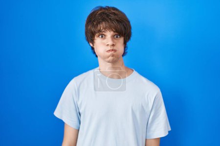 Téléchargez les photos : Hispanic young man standing over blue background puffing cheeks with funny face. mouth inflated with air, crazy expression. - en image libre de droit