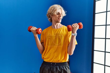 Téléchargez les photos : Jeune homme blond souriant confiant en utilisant des haltères de formation au centre sportif - en image libre de droit