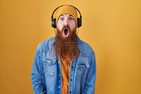 Photo for Caucasian man with long beard listening to music using headphones afraid and shocked with surprise and amazed expression, fear and excited face. - Royalty Free Image