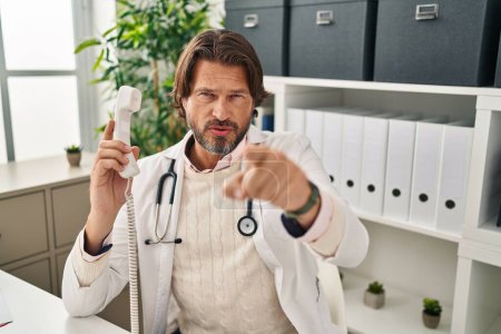 Photo for Handsome middle age doctor man speaking on the phone at the clinic pointing with finger to the camera and to you, confident gesture looking serious - Royalty Free Image