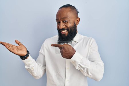 Téléchargez les photos : Afro-Américain debout sur fond bleu étonné et souriant à la caméra tout en présentant avec la main et pointant du doigt. - en image libre de droit