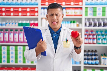 Téléchargez les photos : Jeune homme hispanique travaillant à la pharmacie pharmacie tenant coeur langue collante heureux avec une expression drôle. - en image libre de droit