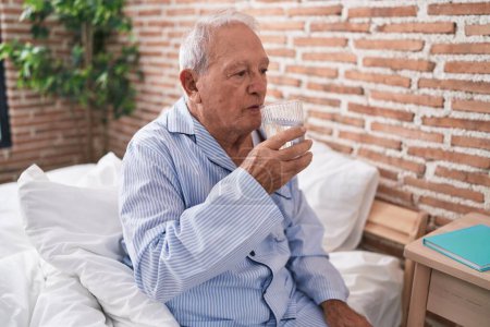 Foto de Hombre de pelo gris de mediana edad bebiendo vaso de agua sentado en la cama en el dormitorio - Imagen libre de derechos