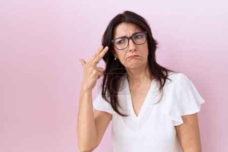 Photo for Middle age hispanic woman wearing casual white t shirt and glasses shooting and killing oneself pointing hand and fingers to head like gun, suicide gesture. - Royalty Free Image