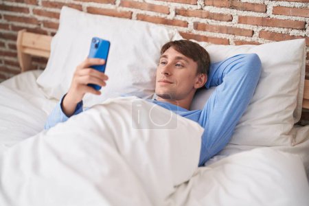 Téléchargez les photos : Jeune homme caucasien utilisant smartphone couché sur le lit dans la chambre - en image libre de droit