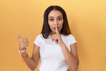 Photo for Young arab woman wearing casual white t shirt over yellow background asking to be quiet with finger on lips pointing with hand to the side. silence and secret concept. - Royalty Free Image