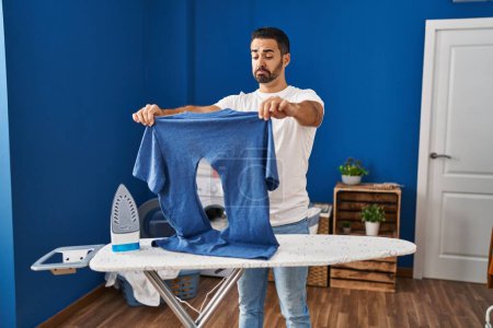 Photo for Young hispanic man with beard ironing holding burned iron shirt at laundry room depressed and worry for distress, crying angry and afraid. sad expression. - Royalty Free Image