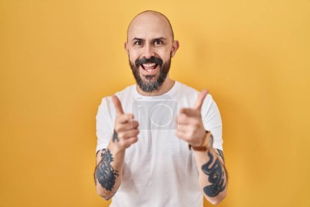 Foto de Joven hombre hispano con tatuajes de pie sobre fondo amarillo señalando con los dedos a la cámara con cara alegre y divertida. buena energía y vibraciones. - Imagen libre de derechos