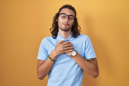 Foto de Young hispanic man standing over yellow background smiling with hands on chest with closed eyes and grateful gesture on face. health concept. - Imagen libre de derechos