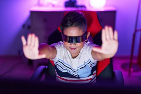 Téléchargez les photos : Adorable garçon hispanique streamer jouer à un jeu vidéo en utilisant des lunettes de réalité virtuelle à la salle de jeu - en image libre de droit