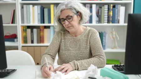 Photo for Middle age woman with grey hair teacher writing on notebook sitting on table at university classroom - Royalty Free Image