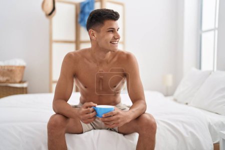 Téléchargez les photos : Jeune homme hispanique buvant une tasse de café assis sur le lit dans la chambre - en image libre de droit