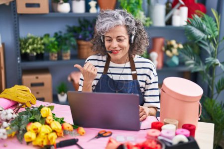Photo for Middle age woman with grey hair working at florist with laptop pointing thumb up to the side smiling happy with open mouth - Royalty Free Image