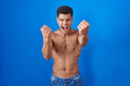 Foto de Joven hombre hispano de pie sin camisa sobre fondo azul enojado y loco levantando puños frustrado y furioso mientras gritaba con ira. rabia y concepto agresivo. - Imagen libre de derechos