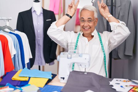 Photo for Middle age man with grey hair dressmaker using sewing machine posing funny and crazy with fingers on head as bunny ears, smiling cheerful - Royalty Free Image