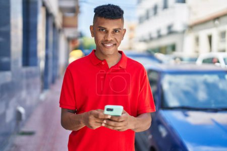 Téléchargez les photos : Jeune homme latino souriant confiant en utilisant smartphone à la rue - en image libre de droit