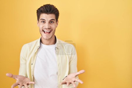 Foto de Young hispanic man standing over yellow background smiling cheerful with open arms as friendly welcome, positive and confident greetings - Imagen libre de derechos