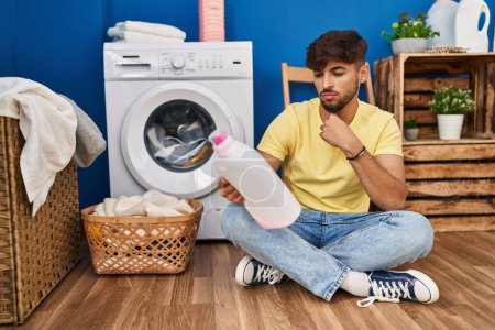 Photo for Young arab man looking detergent bottle sitting on floor at laundry room - Royalty Free Image