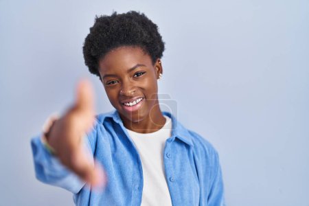 Foto de Mujer afroamericana de pie sobre fondo azul sonriente amistoso ofreciendo apretón de manos como saludo y bienvenida. negocios exitosos. - Imagen libre de derechos
