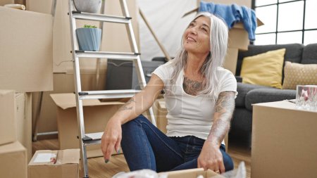 Téléchargez les photos : Moyen âge femme aux cheveux gris souriant confiant assis sur le sol à la nouvelle maison - en image libre de droit