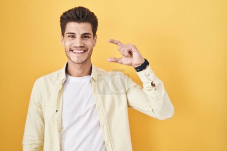 Photo for Young hispanic man standing over yellow background smiling and confident gesturing with hand doing small size sign with fingers looking and the camera. measure concept. - Royalty Free Image