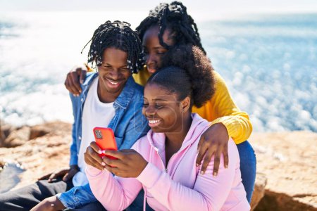 Foto de African american friends using smartphone sitting on rock at seaside - Imagen libre de derechos